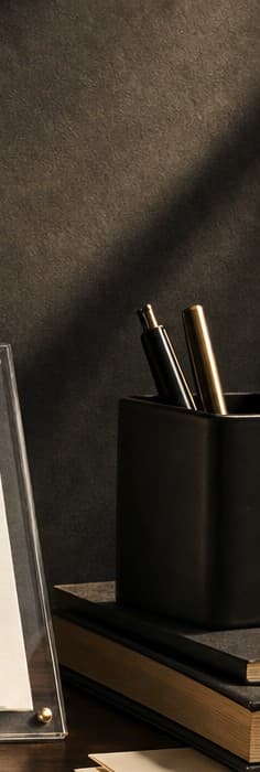 A black pen cup with brass pens beside stacked books under warm light
